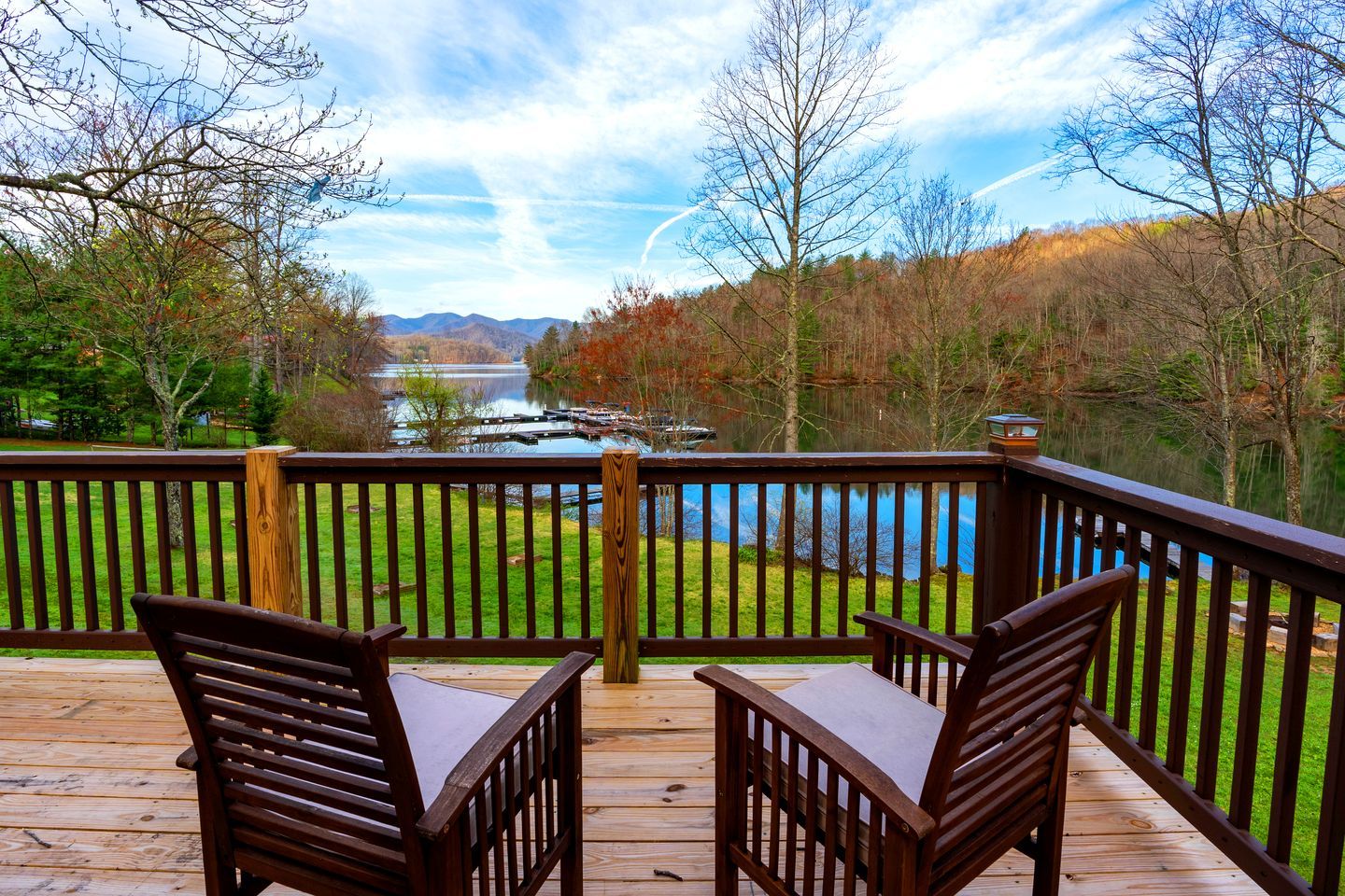 Una terraza de madera con dos sillas y una mesa, con vista a un lago y colinas boscosas.