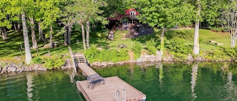 A dock extending into a lake with a wooden platform and a ladder, surrounded by trees and a small building in the background.
