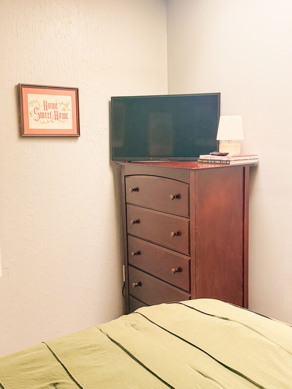 A room with a bed, a dresser with a television on top, a framed sign on the wall, and a lamp on the dresser.