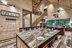 A wooden interior with a dining table set for six, a fireplace, a staircase, and a view of greenery outside. - Fantastic Cabin Nestled Amidst Trees and Hot-tub Provided in Sevierville, Tennessee (Sevierville)