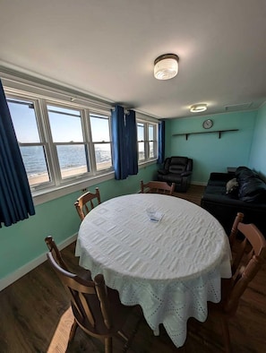 Ein Zimmer mit einem runden Tisch, Stühlen, einer Couch, einem Fenster mit Vorhängen, einer Uhr und Blick aufs Meer.