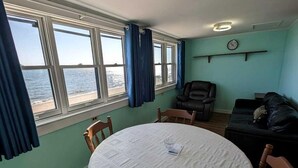 Huoneessa on pyöreä pöytä, tuoleja, sohva, ikkuna, jossa on verhot, kello ja näkymä merelle.