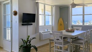 Una habitación con mesa de comedor, sillas, televisor, una tabla de surf y vista al océano.