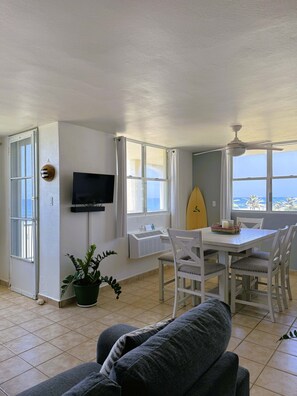 A room with a dining table, chairs, a television, a surfboard, and a view of the ocean. - #31 REAL Oceanfront Second Floor Apt @ Jobos Beach (San Antonio)