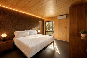 Ein Schlafzimmer mit Holzboden, einem großen Bett, Nachttischlampen und Blick auf Bäume durch ein Fenster.