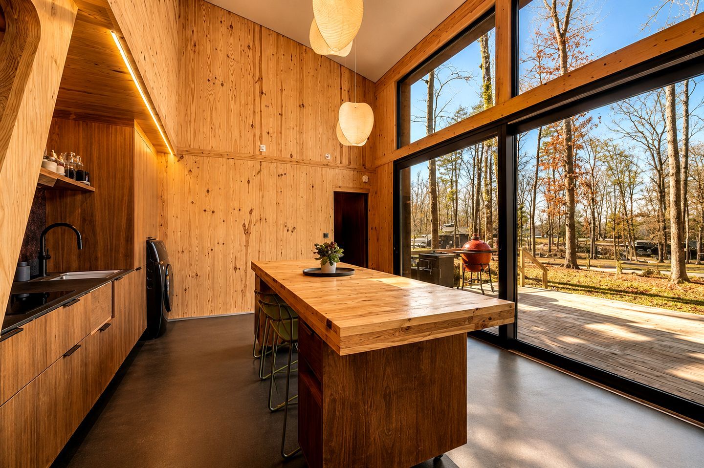 Une cuisine moderne avec des murs en bois, un grand îlot central et une vue sur une zone boisée grâce à de grandes fenêtres.