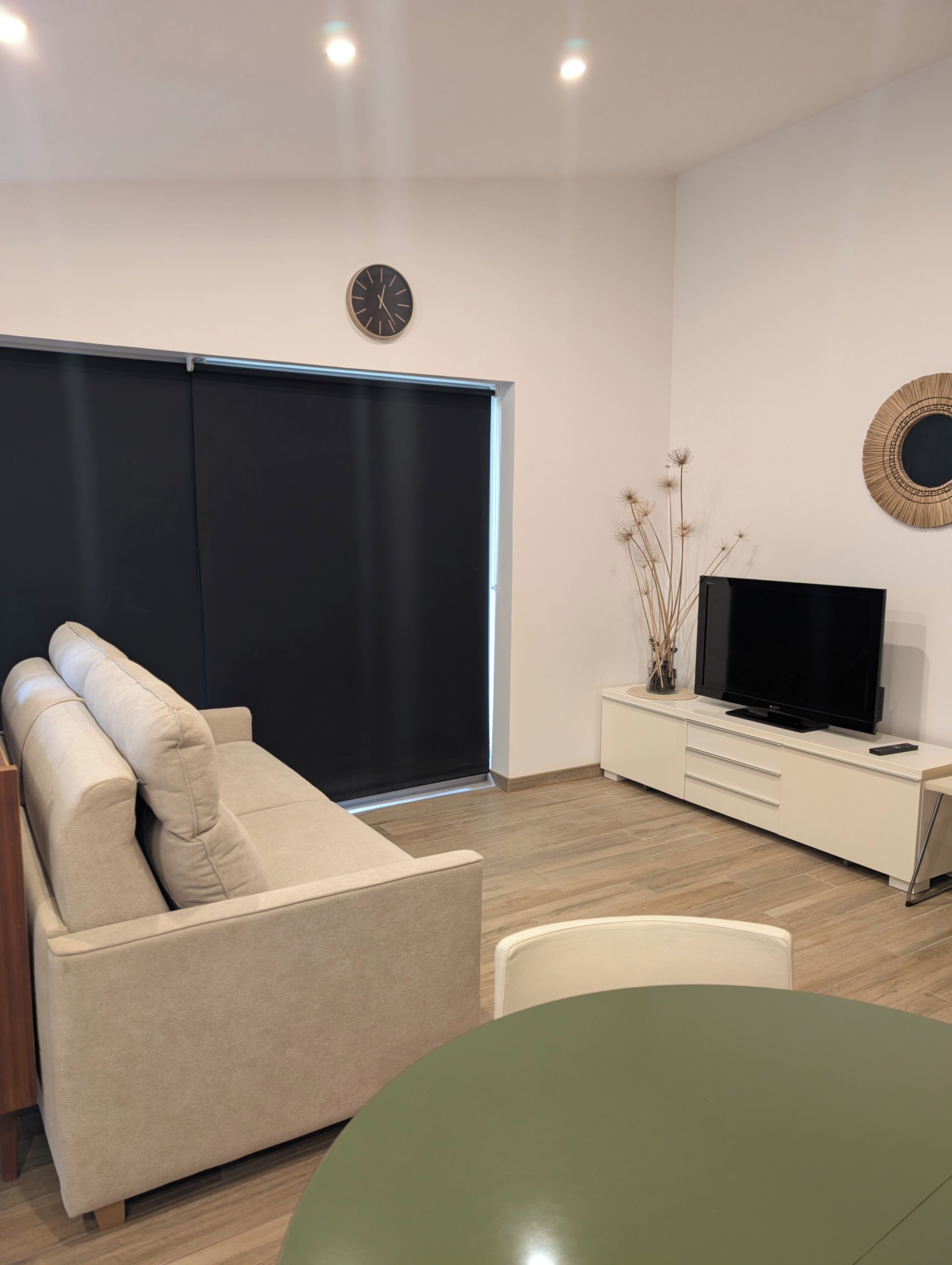 En moderne stue med en sofa, et fjernsyn på et hvidt skab, et rundt bord og et stort vindue med en sort markise.