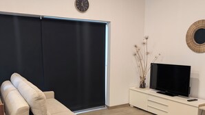 A modern living room with a sofa, a TV on a white cabinet, a round table, and a large window with a black blind. - Le Fournil, a quiet one-room studio near the beaches (Courtils)