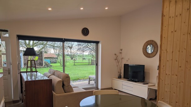 A modern living room with a sofa, a dining table, and a TV on a white cabinet. - Le Fournil, a quiet one-room studio near the beaches (Courtils)