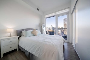 Ein modernes Schlafzimmer mit einem großen Bett, einem Nachttisch und Blick auf eine Stadtlandschaft durch ein großes Fenster.