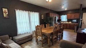 Uma sala de estar moderna com uma mesa de jantar de madeira, sofás, um ventilador de teto e uma área de cozinha ao fundo.