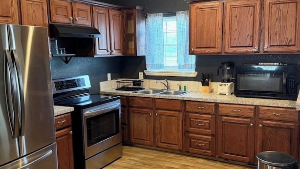 Uma cozinha com armários de madeira, eletrodomésticos de aço inoxidável, uma janela com cortinas de renda e uma lixeira.