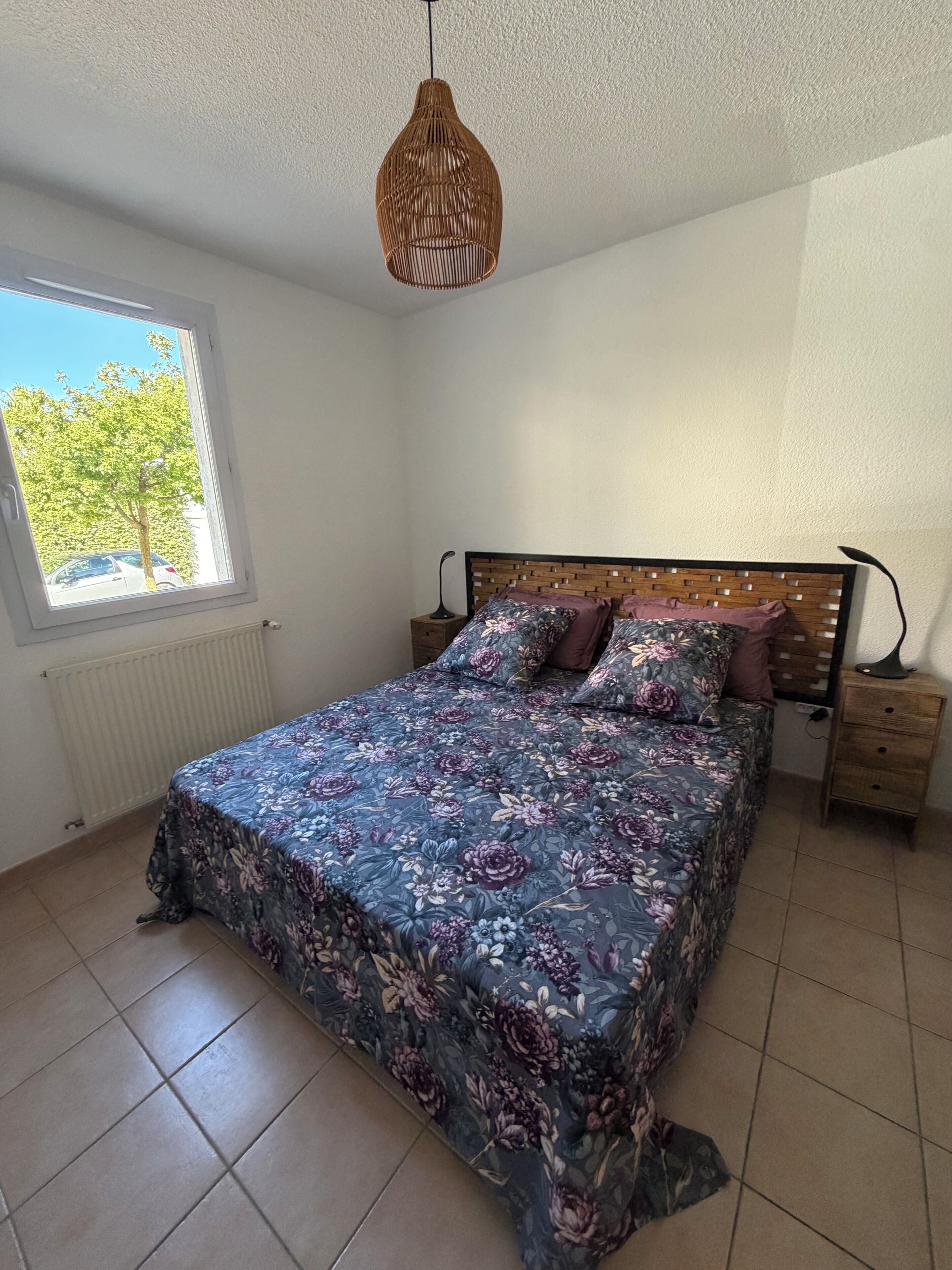 A bedroom with a bed, a nightstand, a lamp, a window, and a hanging light fixture.