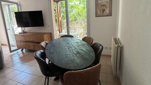 Een eetgedeelte met een ronde tafel, stoelen, een flatscreen televisie en een raam met uitzicht op groen.