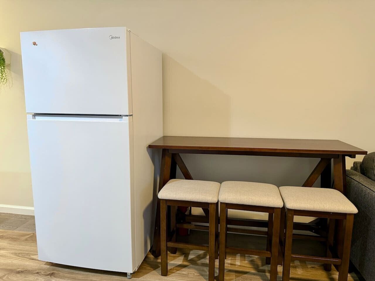 A compact kitchenette with a white refrigerator, a wooden table with two cushioned chairs, and a grey sofa in the background.