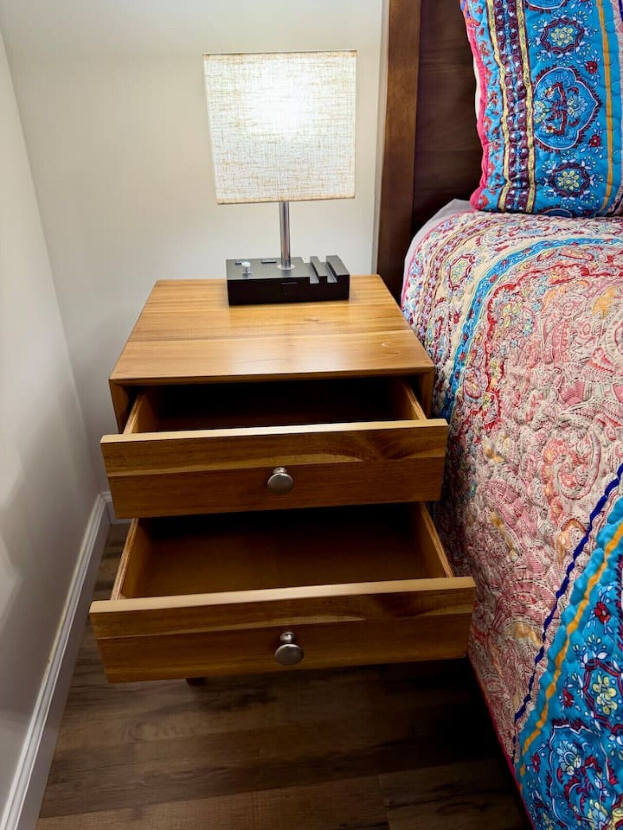 A wooden nightstand with a lamp, two drawers, and a bed with a patterned bedspread.