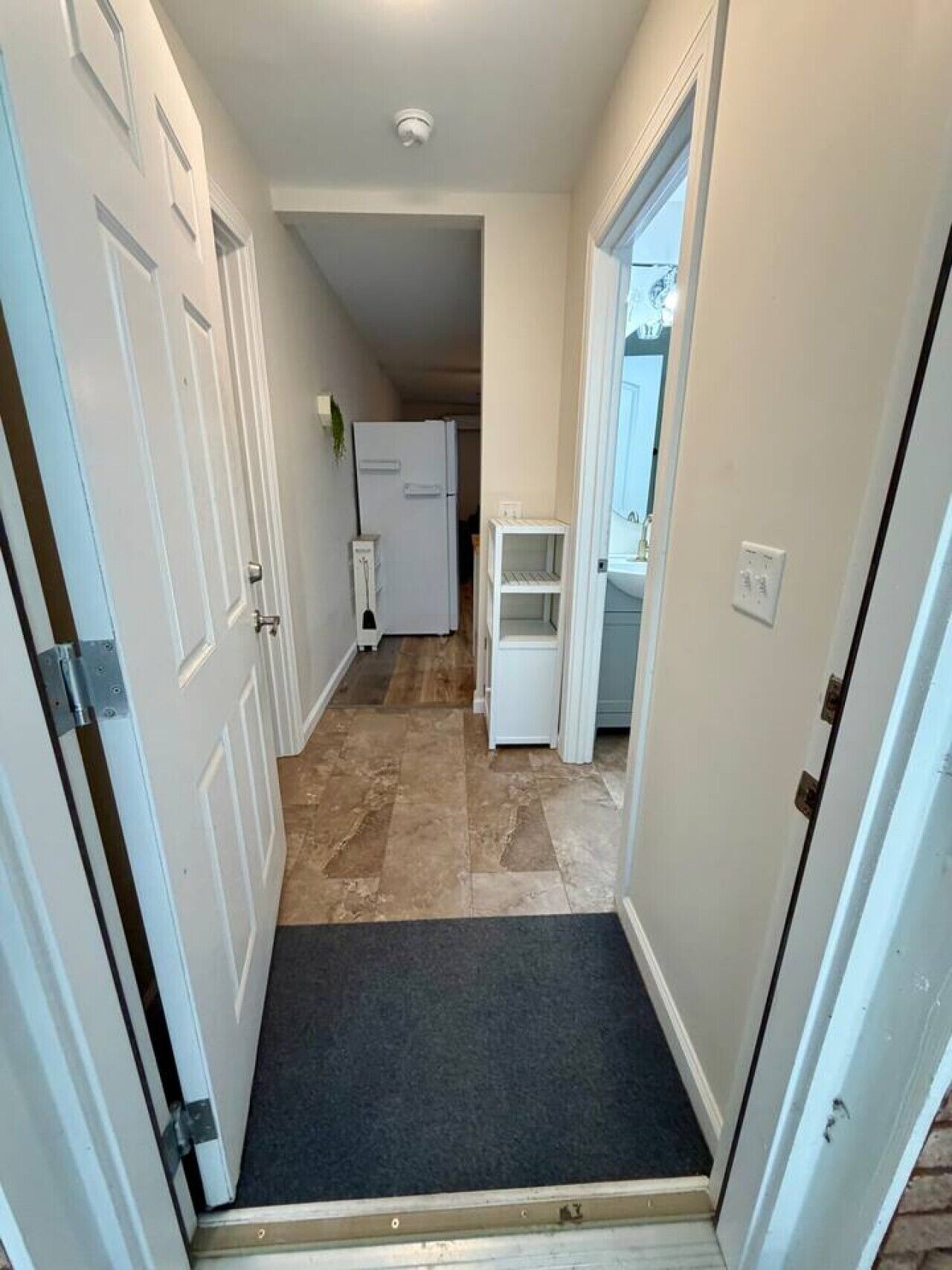 A narrow hallway with white doors, a black mat at the entrance, a white refrigerator, and a small white shelf.
