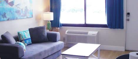 A living room with a sofa, a table, and a chair. There is a window with curtains, a lamp, and an air conditioning unit.