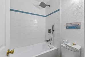 Ein Badezimmer mit weißen Fliesen, einer Duschbrause, einem Toilettenpapierhalter und einem Schild an der Wand.