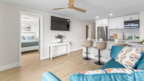 Un salon moderne comprenant un canapé, un téléviseur, un ventilateur de plafond et un coin cuisine avec des placards et un plan de travail.