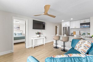 Ein modernes Wohnzimmer mit einer Couch, einem Fernseher, einem Deckenventilator und einer Küchenzeile mit Schränken und einer Arbeitsplatte.