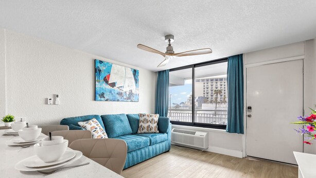 Un salon moderne avec un canapé bleu, une table à manger avec de l’assiette blanche, un ventilateur de plafond et une grande fenêtre avec des rideaux.