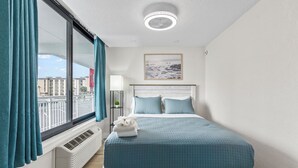 Une chambre avec une grande fenêtre, un lit avec une literie bleue, un ventilateur de plafond et un tableau encadré au mur.