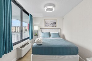 Ein Schlafzimmer mit einem großen Fenster, einem Bett mit blauer Bettwäsche, einem Deckenventilator und einem gerahmten Bild an der Wand.