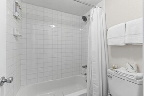 Kylpyhuone, jossa on valkoiset laatat, kylpyamme, suihkupää, wc ja pyyhekoukku.