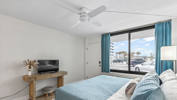 Ein Schlafzimmer mit Bett, Fernseher auf Holzständer, Deckenventilator, Fenster mit Vorhängen und Blick auf die Straße.