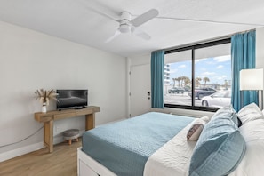 Ein Schlafzimmer mit Bett, Fernseher auf Holzständer, Deckenventilator, Fenster mit Vorhängen und Blick auf die Straße.