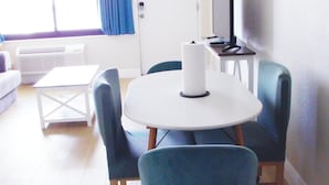 Una stanza con un tavolo rotondo, sedie blu, un divano bianco, un televisore su un mobile e una finestra con tende.