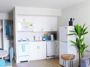 A compact kitchen with white cabinets, a microwave, a stove, and a dishwasher. There is a dining area with a table and chairs, and a potted plant. - Direct Oceanfront Daytona Beach Condo | Full Kitchen | Beach Access (Daytona Beach)
