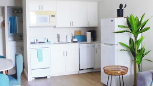 Una cucina compatta con mobili bianchi, un forno a microonde, un fornello e una lavastoviglie. C'è un'area pranzo con tavolo e sedie, e una pianta in vaso.