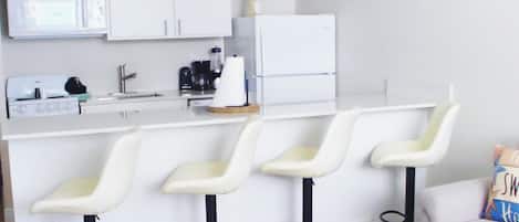 A modern kitchen with white cabinets, a microwave, a coffee maker, and a refrigerator. There are three bar stools with white seats and black bases.