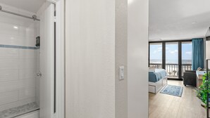 A modern hotel room with a shower, a balcony with a view, a desk, and a bed. - Direct Oceanview Daytona Beach Condo | Full Kitchen | Beach Access (Daytona Beach Shore)