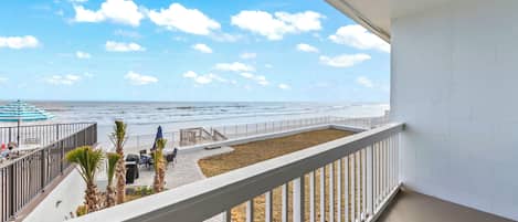 Un balcón con vistas a la playa, un área de estar con mesa y sillas, y un pequeño jardín con palmeras.
