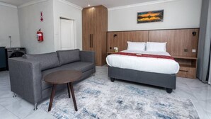 A hotel room with a grey sofa, a round wooden table, a bed with a white and red blanket, a wooden headboard, a painting on the wall, and a fire extinguisher.