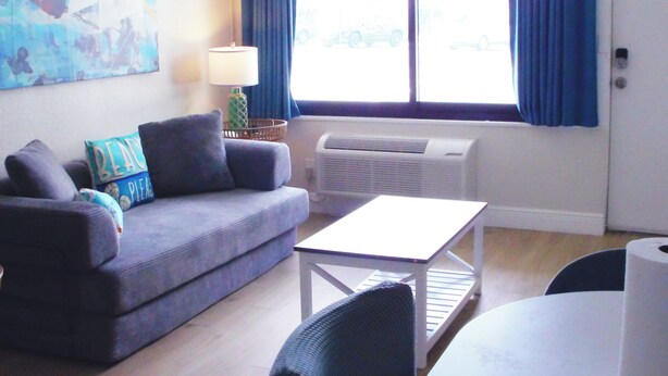 A living room with a sofa, a table, and a chair. There is a window with curtains, a lamp, and an air conditioning unit. - Direct Oceanfront Daytona Beach Condo | Full Kitchen | Beach Access (Daytona Beach)