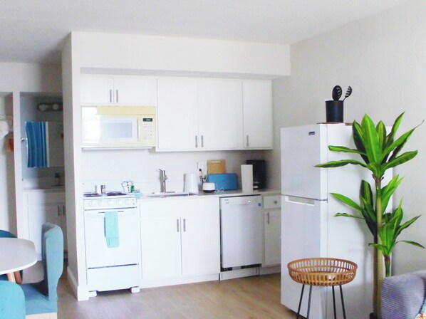 Una cucina compatta con mobili bianchi, un forno a microonde, un fornello e una lavastoviglie. È presente una zona pranzo con tavolo e sedie. Si nota una pianta nell'angolo.