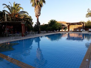 A swimming pool with lounge chairs, palm trees, and a building with a tiled roof. - 2BR Family Apartment in Gouvia – Apt 2 (Kontokali)