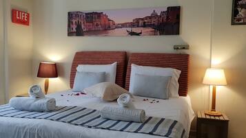 A hotel room with a bed, two bedside lamps, a painting of a canal scene, and a 'LIFE' sign on the wall.