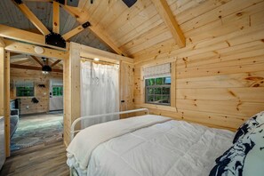A wooden cabin interior with a bed, a window with a view, a door leading to another room, and a ceiling fan. - Charming Forest Cabin with Hot Tub & Covered Porch in Mentone, Alabama (Mentone)