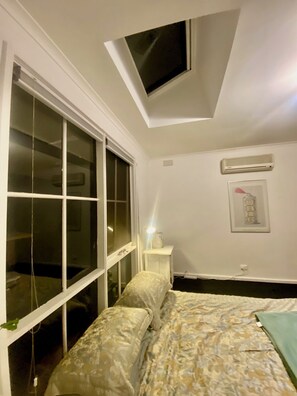 A bedroom with a skylight, a bed with patterned bedding, a bedside table with a lamp, and a wall-mounted air conditioner. - Funky house close to Castlemaine, surrounded by extensive gardens and bushland. (Golden Point)
