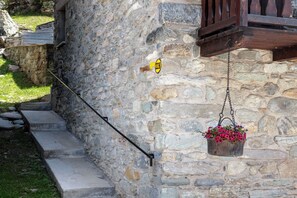Ein Steinhaus mit einer Steintreppe, einem hängenden Blumenkorb und einem gelben Schild mit schwarzem Pfeil.
