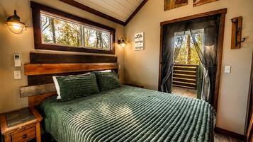 A bedroom with a wooden headboard, a green textured bedspread, a nightstand, a wall-mounted light, and a window with a view of trees.
