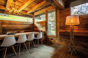 Ein Holzinnenraum mit Esstisch, Stühlen, einer Deckenleuchte, einer Lampe und großen Fenstern.