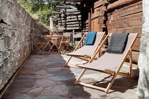 Une terrasse dallée de pierres, agrémentée de chaises longues en bois et d’une table, le tout entouré de murs en bois rustique.