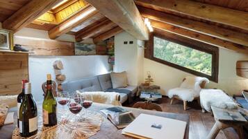 A cozy attic room with wooden beams, a loft bed, a sofa, a dining table with wine bottles and glasses, and a view of greenery outside.