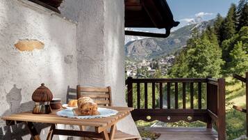 A wooden table with a rustic chair, a bread loaf, and a coffee pot on a balcony overlooking a mountainous town.